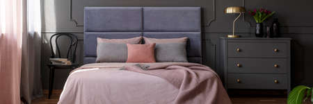 Pastel pink bedclothes on king-size bed with soft bedhead standing in dark bedroom interior with gold lamp and fresh tulips on grey cupboardの写真素材