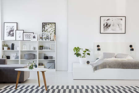 Real photo of a platform bed standing next to a sofa and a table in a one room flat interior with...