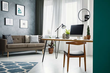 Wooden chair at desk in living room interior with work area and posters on grey wall. Real photoの写真素材