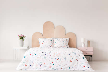 Patterned bed with headboard between table with flowers and pink cabinet in bedroom interior. Real...