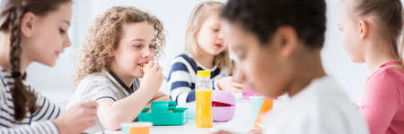 Panorama of school kids eating lunch together during a break, at a school canteen. Focus on a boy with curly hair eating a sandwichの写真素材