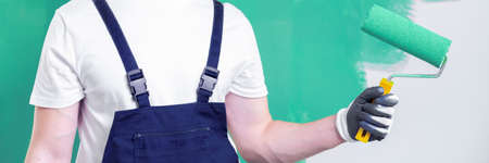 Close-up on a home renovation worker in workwear holding a roller with a neo mint paint and a work-in-progress, green wall in the backgroundの写真素材