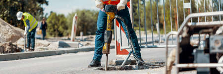 Builder drilling the road with a tool and his coworker at the city road work construction site in the blurry backgroundの写真素材