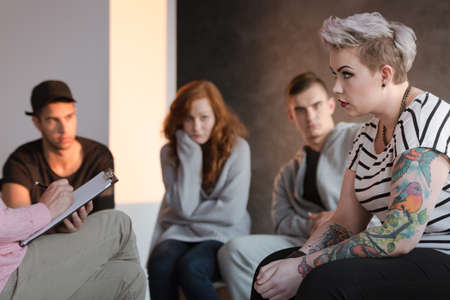 Tattooed rebel teenage girl sitting in front of a therapist who is taking notes during a group psychotherapy meeting. Other young people in the blurred background.の写真素材
