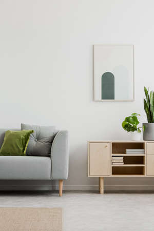 Poster above cupboard in simple living room interior with grey settee and plants. Real photo
