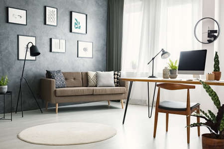 Chair at desk with desktop computer in grey open space interior with brown sofa under posters. Real...