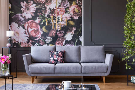 Grey lounge with patterned cushion in real photo of dark living room interior with floral wallpaper,...