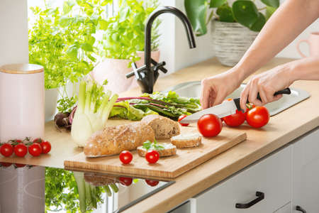 Woman preparing healthy breakfast, cutting a tomato in half and organic herbs and vegetables in a...