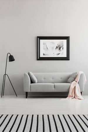 Poster above grey sofa with pink blanket in living room interior with lamp and striped carpet. Real...
