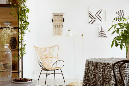 Real photo of a garden chair standing against white wall with macrame and monochromatic paintings in...