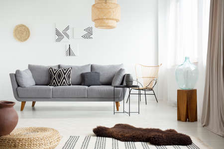 Real photo of a brown, fur rug lying on white floor in front of a gray couch with cushions in bright...