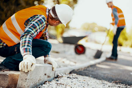 Worker with gloves and in a helmet arranging curbs on the street. Blurred man in the backgroundの写真素材