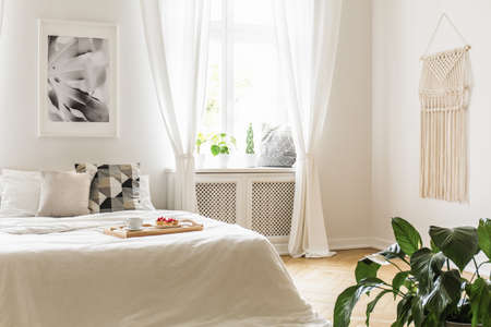 Breakfast tray with pastries and coffee on a cozy, white bed in a bright and peaceful bedroom interior with plants on a window sill seatの写真素材