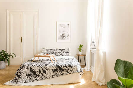Comfortable big bed with white and black flower design bedding and a breakfast tray on in a wooden floor bright bedroom interior. Double door to another room in a background wall. Real photo.の写真素材