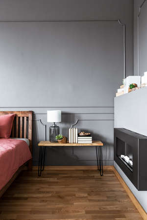 Lamp on wooden table between pink bed and black fireplace in grey cozy bedroom interiorの写真素材