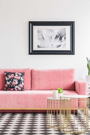 Velvet couch with a floral pillow, poster on the wall and metal coffee tables in a living room interior with checkered floor. Real photoの写真素材
