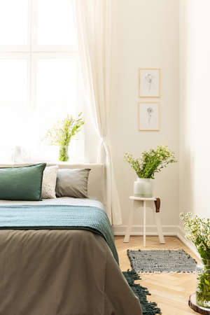 Posters on white wall above flowers on table next to grey and green bed in bedroom interior. Real photoの写真素材