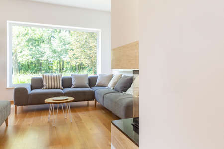 Pillows on grey corner couch in bright living room interior with window and wooden table. Real photo...