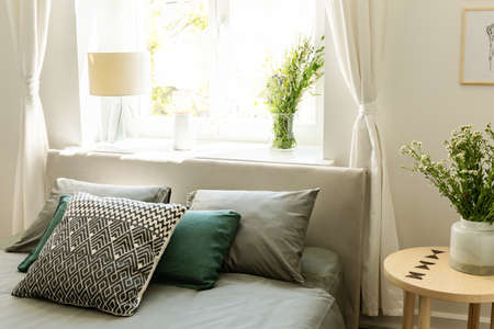 Patterned green and grey pillows on bed next to table with flowers in bedroom interior. Real photoの写真素材