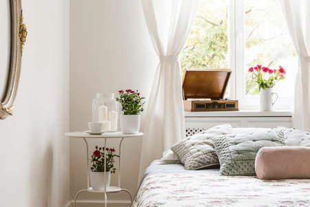 White bedroom interior in real photo with bedside table with tea cup, candles and red roses, windowsill with gramophone and fresh flowersの写真素材