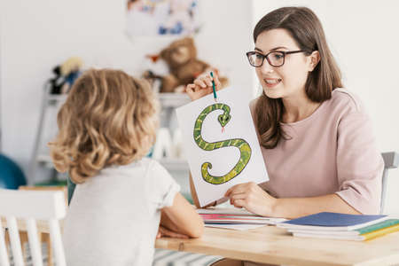 A child with development problems with a professional speech therapist during a meeting. Tutor holding a prop poster of a snake as a letter 's'.