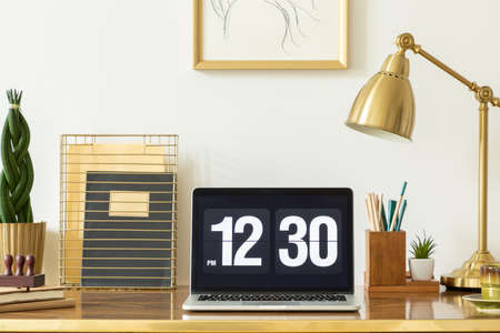 Laptop showing the time, notebooks, plant and lamp on a desk in an office interior. Real photoの写真素材