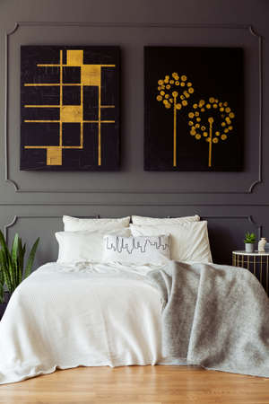 Black and gold posters above white bed with blanket in grey bedroom interior. Real photo