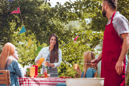A young girl pouring orange juice into a cup and a man in a burgundy apron grilling during a...