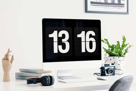 Computer screen showing time, headphones, camera, books, wooden hand an plant on a desk in a bright...