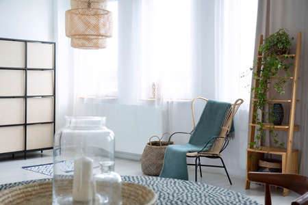 Green blanket on armchair in bright living room interior with plant, lamp and windows. Real photo