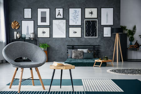 Grey armchair next to table on carpet in living room interior with gallery of posters. Real photo with blurred backgroundの写真素材