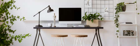 Black desk with lamp, tea cup, notebooks and mockup screen in real photo of bright living room interior with fresh plantsの写真素材