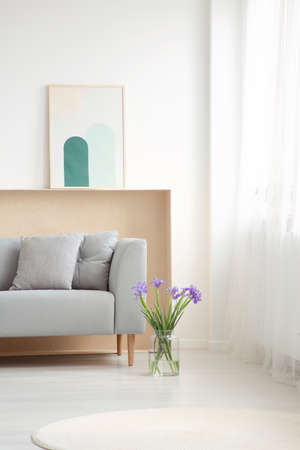 Blue flowers next to grey couch with cushions in simple living room interior with poster. Real photoの写真素材