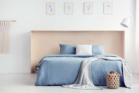 Blanket and pillows on blue bed in white minimal bedroom interior with posters and lamp. Real photoの写真素材