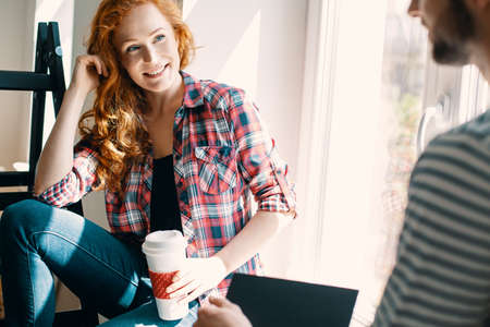 Smiling red haired woman drinking coffee and talking with friend at homeの写真素材