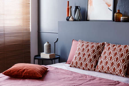 Patterned cushions on bed with headboard in grey modern bedroom interior with blindsの写真素材