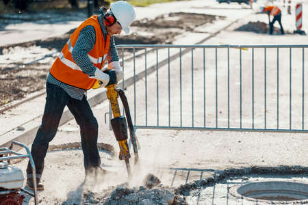 Craftsman in reflective vest and white helmet while using drill on the streetの写真素材