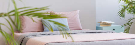 Real photo with plant in blurred foreground of bright bedroom interior with books on bedside table...
