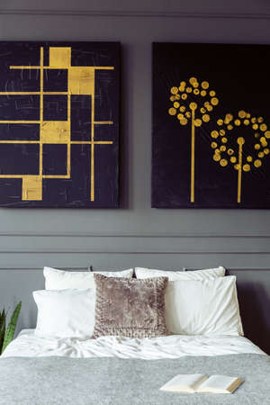 Real photo of a bedroom interior with black and gold paintings, double bed with pillows and book