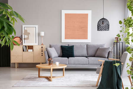 Pink poster above grey sofa in living room interior with wooden table near cupboard. Real photo