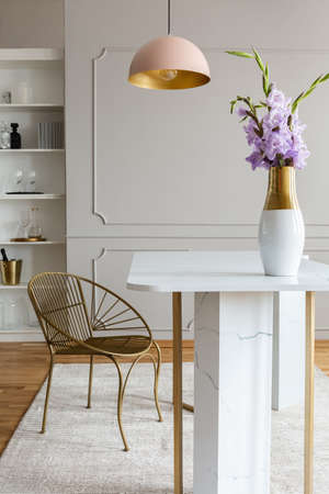 Flowers on white marble table near gold chair under pink lamp in grey dining room interior. Real...