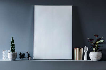 Real photo of a mockup poster standing on shelf between plants and ornaments in grey room interior....
