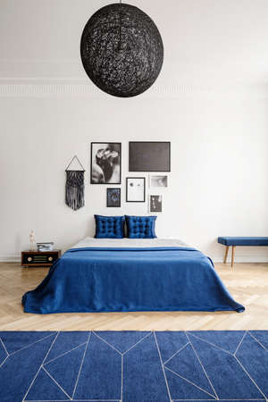 Lamp above blue bed with pillows in white bedroom interior with posters and carpet. Real photo