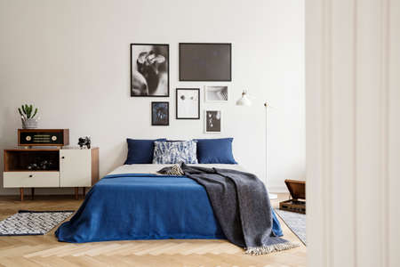 Blanket on blue bed with pillows in retro bedroom interior with radio on cabinet and posters. Real...