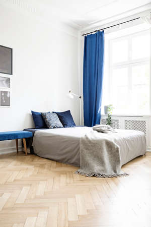 Grey blanket on bed in bright bedroom interior with lamp and navy blue drapes at window. Real photoの写真素材