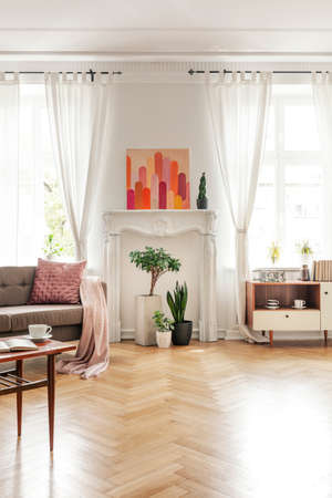Wooden table and beige couch in spacious living room interior with poster and plants. Real photo