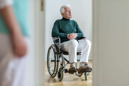 Sad disabled elderly man in a wheelchair in the hospital. Blurred nurse in a foreground