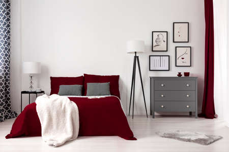 Real photo of a glamour bedroom interior with dark red bed, blanket, lamp and grey commode. Empty...