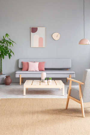 Wooden table between armchair and grey sofa in apartment interior with painting and carpet. Real photoの写真素材