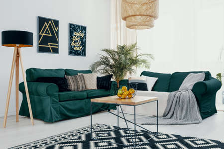 Wooden table in front of green sofa in white living room interior with lamp and posters. Real photo